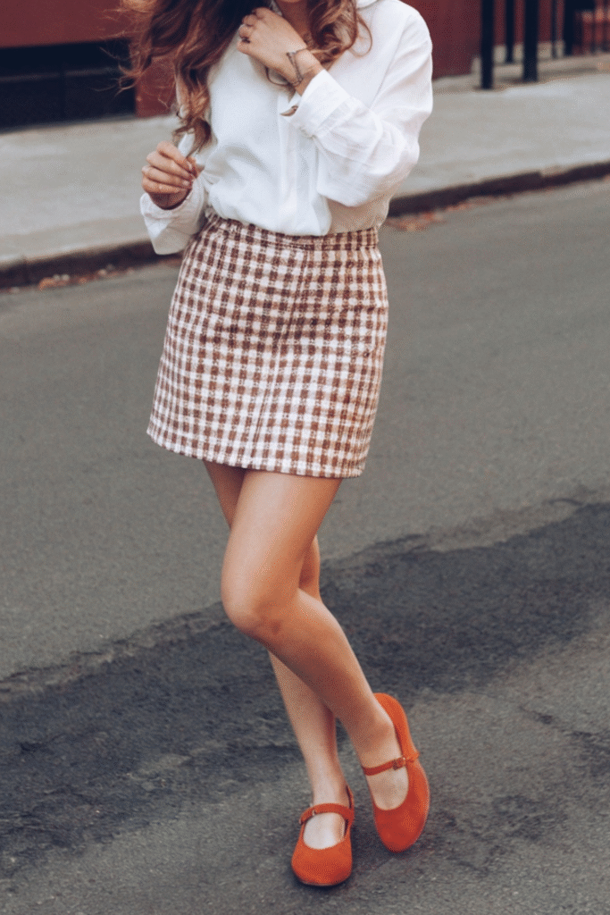 Woman in a brown gingham mini skirt and white blouse, smiling while walking down a city street in orange Mary Jane shoes.