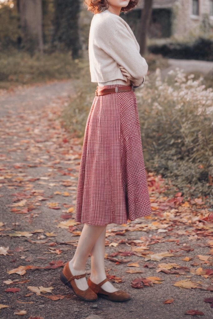 Woman wearing a sleeveless plaid gingham dress in muted autumn tones, cinched with a tan leather belt, paired with black tights and heeled Mary Janes, perfect for vintage-inspired fall style.