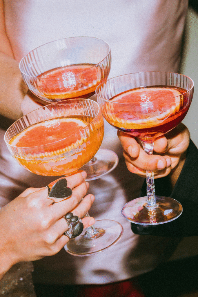 Close-up of three hands holding coupe glasses filled with grapefruit-garnished cocktails, perfect for a stylish Friendsgiving celebration