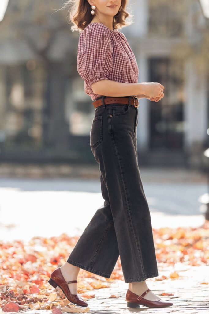 Woman in puff-sleeve gingham blouse with black wide-leg jeans and brown Mary Janes walking through autumn leaves.