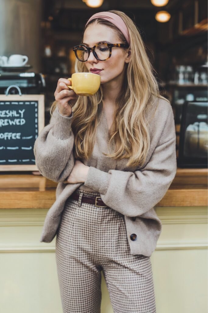 Woman in taupe cardigan and brown gingham pants sipping coffee indoors for a cozy vintage café style.