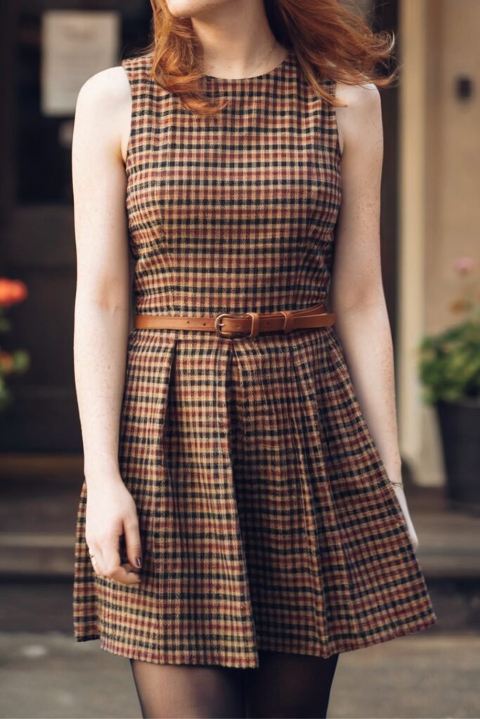 Woman wearing a sleeveless vintage-inspired plaid gingham fit-and-flare dress with a brown belt, styled for fall. The checkered pattern combines warm autumn tones, creating a timeless day-to-night look.